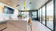 Located at 2141 Waterside Drive in Nashville, TN, this contemporary kitchen space delivers a polished, high-design aesthetic with an impressive array of upscale finishes. At the heart of the room sits a commanding marble island with dramatic veining, complemented by stylish wood-and-brass bar stools that invite casual seating and social gatherings. Overhead, a sculptural black-and-gold multi-arm pendant light fixture serves as a bold artistic focal point, adding warmth and visual drama to the space. The kitchen wall features a textured white geometric tile backsplash, dark cabinetry with gold hardware, a stainless steel under-counter refrigerator, and a wall-mounted flat-screen television. Light hardwood flooring runs throughout, enhancing the bright and airy atmosphere. Floor-to-ceiling windows and a glass-panel sliding door flood the interior with abundant natural light while framing sweeping views of the Nashville skyline. The sliding door opens directly to an outdoor terrace furnished for relaxation, seamlessly blending indoor and outdoor living. Charcoal-toned walls provide a sophisticated contrast to the bright finishes, creating a refined and cohesive modern ambiance throughout the space.