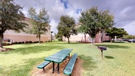This outdoor communal space is situated within a multi-building residential community, offering a welcoming and functional gathering area for residents. The centerpiece of the space is a generously sized, well-kept lawn framed by mature shade trees, including oaks and palms, which provide a natural canopy and a sense of established character. The open grassy area creates a spacious feel, ideal for leisurely outdoor activities or casual gatherings. A green metal picnic table with attached bench seating is positioned on the lawn, offering a designated spot for outdoor dining or socializing. Adjacent to the seating area, a charcoal grill on a post-mounted stand provides a convenient amenity for outdoor cooking and entertaining. The surrounding multi-story buildings, finished in warm earth tones, enclose the courtyard on multiple sides, contributing to a sense of privacy and community. The sky above is open and expansive, allowing ample natural light to illuminate the space throughout the day. Overall, the courtyard exudes a relaxed, park-like ambiance, making it a practical and pleasant outdoor retreat within the community.
