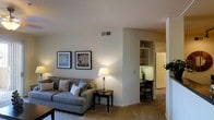 This single-floor residence offers a thoughtfully arranged layout designed for comfortable everyday living. The living area serves as the heart of the home, featuring soft carpeted flooring and warm neutral wall tones that create an inviting, relaxed ambiance. Tasteful framed artwork adorns the walls, complementing the understated décor throughout. A stylish table lamp provides warm accent lighting, enhancing the cozy atmosphere of the space.

Adjacent to the living area, a built-in shelving niche offers practical display and storage space, adding a subtle architectural detail to the open flow of the floor plan. Beyond, a compact dining nook accommodates a small table and chairs, making efficient use of the available space.

A hallway extends further into the residence, lined with track lighting overhead and decorated with a decorative round wall accent, leading to additional rooms. The overall layout benefits from a smooth, open transition between living, dining, and connecting spaces, giving the home a cohesive and functional feel. The consistent use of neutral finishes, carpet flooring, and warm artificial lighting throughout lends the space a unified, welcoming character well-suited for both relaxing and entertaining.