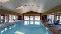 This single-story indoor pool facility offers a welcoming and functional aquatic environment designed for both recreation and relaxation. The centerpiece of the space is a generously sized rectangular swimming pool with clear, blue water, providing ample room for lap swimming and leisure. The pool deck is finished in light-toned tile and is lined with blue lounge chairs on both sides, creating a comfortable area for guests to unwind.

The architectural design is highlighted by a vaulted, cathedral-style ceiling clad in light wood paneling, which lends the space a warm and airy character. A pair of skylights are set into the ceiling, allowing natural light to filter in and brighten the interior throughout the day. The walls are finished in a rich wood tone complemented by brick accents, reinforcing a classic and inviting aesthetic.

A row of large windows and glass-paneled doors runs along the far wall, offering views of the outdoor grounds and flooding the space with additional natural light. The doors provide convenient access to the exterior area. Utility features such as ventilation grilles are neatly integrated into the ceiling. Overall, the space balances practical amenity with a comfortable, resort-like ambiance suited to a variety of users.