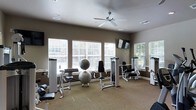 This fitness center offers a thoughtfully designed workout environment spread across a single floor. The space is finished with neutral, warm-toned walls and carpeted flooring, creating a comfortable and inviting atmosphere for exercise. Large windows fitted with white horizontal blinds line multiple walls, allowing an abundance of natural light to fill the room while providing views of the surrounding greenery outdoors. A ceiling fan centered overhead ensures comfortable air circulation throughout workouts. The room is equipped with a range of strength-training machines, including cable and resistance equipment suited for both upper and lower body exercises. Stability balls and additional accessories complement the machine offerings, catering to a variety of fitness routines. A wall-mounted flat-screen television adds an entertainment element, making workouts more engaging. Recessed lighting supplements the natural light for evening use. The overall layout is open and organized, providing ample space to move between equipment comfortably. The clean, modern aesthetic and practical amenities make this fitness center a functional and welcoming space for users of all fitness levels.