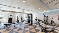 Located at 1230 Front Pl in North Port, Florida, this fitness center offers a generously proportioned, single-floor exercise space designed to support a comprehensive workout experience. The room makes a strong visual impression with its bold geometric-patterned flooring in neutral tones of tan, beige, and dark brown, lending a modern, dynamic aesthetic throughout.

The ceiling is a standout architectural feature, showcasing a textured diamond-pattern tray design with recessed lighting that illuminates the space evenly. Large ceiling fans — including an oversized multi-blade fan — ensure excellent air circulation, keeping the environment comfortable during intense workouts. Windows along the perimeter allow natural light to filter in, contributing to an open, airy ambiance.

The fitness floor is comprehensively equipped with a wide range of cardio and strength training machines, including cable systems, stair climbers, stationary bikes, treadmills, and rowing equipment. A dedicated free weights section features a full dumbbell rack alongside large wall-mounted mirrors and flat-screen televisions. Built-in shelving units and cabinetry provide organized storage, while motivational wall graphics reinforce an energetic atmosphere. French doors offer easy access, completing a fitness facility that is both functional and thoughtfully designed.
