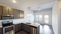 This single-floor unit showcases a thoughtfully designed open-concept layout with consistent modern finishes throughout. The kitchen is a standout feature, equipped with sleek gray cabinetry extending to the ceiling, granite countertops, and a full suite of stainless steel appliances including a range, over-the-range microwave, dishwasher, and refrigerator. A white subway tile backsplash adds a clean, contemporary touch, while a peninsula with a undermount sink and curved granite edge provides additional prep space and casual seating potential. Pendant lighting hangs above the peninsula, complementing the ceiling fan in the adjacent living area, which receives natural light through windows overlooking an outdoor space. Dark hardwood-style flooring runs throughout the main living and kitchen areas, creating a cohesive and polished aesthetic. The bathroom features matching gray cabinetry with granite countertops, a tiled shower-tub combination, and the same hardwood-style flooring for continuity. A carpeted bedroom offers a comfortable retreat with a reach-in closet providing organized storage. Crown molding details and clean white walls contribute to the overall refined and move-in-ready character of the space.