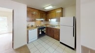 This single-floor space offers a practical and functional layout, with distinct zones that flow naturally into one another. The kitchen is outfitted with warm-toned wood cabinetry arranged in an L-shaped configuration, providing ample upper and lower storage. It includes a black electric range, a white dishwasher, a white refrigerator, and a double basin sink positioned beneath under-cabinet lighting. Light tile flooring extends throughout the kitchen area, offering a clean, easy-to-maintain surface.

The kitchen opens directly into the main living area, which transitions to neutral wall-to-wall carpeting. A ceiling fan with an integrated light fixture is centrally mounted, providing both air circulation and ambient lighting. A window along the far wall allows natural light into the space, and a wall-mounted HVAC unit offers additional climate control. A standard entry door is positioned nearby, providing access to the exterior.

A separate bedroom is carpeted and similarly equipped with a ceiling fan. A large window fitted with blinds allows ample natural light, and tile flooring at the entry transitions smoothly to carpet within the room.

The bathroom features a single-sink vanity with a large mirror, and a full-sized soaking tub with an overhead shower fixture, completing this straightforward and well-organized living space.