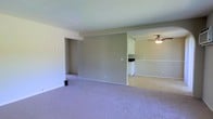 This single-floor space features an open and functional layout designed to maximize usable living area. The main living space is finished with neutral-toned wall-to-wall carpeting, contributing to a warm and understated ambiance. Walls are painted in a soft greige tone, providing a versatile backdrop that complements a variety of interior styles.

A partial dividing wall separates the living area from the adjoining dining or secondary living zone, creating a sense of defined space while maintaining an open feel throughout. This connected area features a ceiling fan with a light fixture, offering both ventilation and ambient lighting.

The kitchen is partially visible beyond the dividing wall and is outfitted with white cabinetry and appears to include standard appliances. The kitchen space transitions naturally from the adjoining area, supporting an easy flow between cooking and living zones.

A recessed nook or alcove is visible along one wall of the main living area, offering potential for additional storage or a designated use. The overall design is clean and straightforward, with consistent flooring and neutral finishes that give the space a move-in ready appearance.