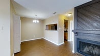 This single-floor residence offers a thoughtfully designed open-concept layout that promotes a seamless flow between living and dining areas. The dining space is anchored by an elegant chandelier overhead, adding a touch of refined character to the room. Warm hardwood-style flooring extends throughout the main living areas, providing both visual warmth and durability.

A defining architectural feature is the pass-through window connecting the dining area to the kitchen, enhancing the open feel while maintaining a sense of distinct zones. The kitchen is outfitted with warm wood cabinetry, dark countertops, and stainless steel appliances, offering a practical and well-appointed cooking space. A ceiling-mounted light fixture illuminates the kitchen, complementing the natural warmth of the cabinetry tones.

The living area features a striking dark-framed fireplace, serving as a bold focal point that grounds the space and adds a cozy ambiance. Neutral beige walls throughout create a clean, versatile backdrop suitable for a variety of décor styles. A textured ceiling adds a subtle design detail visible across the entire space. The layout is efficient and comfortable, making excellent use of the single-floor footprint to deliver a connected and inviting home environment.