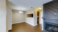 This single-floor residence offers a thoughtfully designed open-concept layout that promotes a seamless flow between living and dining areas. The dining space is anchored by an elegant chandelier overhead, adding a touch of refined character to the room. Warm hardwood-style flooring extends throughout the main living areas, providing both visual warmth and durability.

A defining architectural feature is the pass-through window connecting the dining area to the kitchen, enhancing the open feel while maintaining a sense of distinct zones. The kitchen is outfitted with warm wood cabinetry, dark countertops, and stainless steel appliances, offering a practical and well-appointed cooking space. A ceiling-mounted light fixture illuminates the kitchen, complementing the natural warmth of the cabinetry tones.

The living area features a striking dark-framed fireplace, serving as a bold focal point that grounds the space and adds a cozy ambiance. Neutral beige walls throughout create a clean, versatile backdrop suitable for a variety of décor styles. A textured ceiling adds a subtle design detail visible across the entire space. The layout is efficient and comfortable, making excellent use of the single-floor footprint to deliver a connected and inviting home environment.