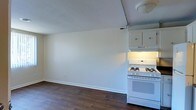 This single-floor residence offers a clean, straightforward layout designed for comfortable everyday living. The interior is characterized by smooth white walls and rich dark hardwood flooring that runs throughout, lending a warm and grounded feel to the space. The kitchen area is efficiently appointed with white cabinetry offering ample overhead storage, a white gas range with a built-in overhead range hood for ventilation, and a bright, utilitarian aesthetic. The kitchen transitions naturally into the adjacent living area, reflecting an open-concept sensibility that allows the space to feel connected and versatile. Natural light fills the room, bouncing off the light-colored walls and ceilings to create a bright, airy ambiance. The overall design is simple and functional, with clean lines and neutral tones that provide a neutral canvas easily personalized to individual taste. The layout is practical, with each area of the home flowing seamlessly into the next, making the most of the available footprint. The combination of dark flooring and white finishes creates a classic contrast that gives the interior a timeless, balanced character.