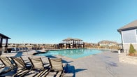 This expansive outdoor amenity space is designed for leisure and community gathering, centered around a generously sized rectangular swimming pool with clear, blue-green water. The pool deck is finished in large-format tile, providing ample space for multiple lounge chairs positioned to take advantage of the open sky and abundant natural sunlight throughout the day.

A dedicated spa is situated adjacent to the main pool, offering a distinct relaxation area. A prominent pergola structure, supported by stacked stone columns, extends across one end of the deck and provides partial shade. Beneath the pergola, additional seating includes lounge chairs and outdoor dining sets with striped market umbrellas, creating comfortable gathering spaces.

The perimeter of the deck is enclosed by a wrought iron fence, beyond which a scenic pond or lake is visible, adding a tranquil waterfront backdrop to the setting. A multi-story clubhouse or amenity building with large windows and a pitched roof anchors the background of the pool area, lending architectural context to the space.

Surrounding residential buildings are visible in the distance, indicating this is a shared community amenity. The overall ambiance is open, bright, and resort-inspired, designed to maximize outdoor enjoyment.