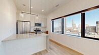 Located at 345 Saint Peter Street in the heart of downtown Saint Paul, MN, this modern studio offers a thoughtfully designed single-floor layout that maximizes both function and style. The open-plan kitchen and living area serves as the centerpiece of the space, featuring light hardwood flooring throughout, crisp white walls, and recessed lighting that keeps the interior bright and airy. The kitchen is outfitted with stainless steel appliances, including a full-size refrigerator, range, and microwave, complemented by a two-tone cabinet design pairing white upper cabinets with warm wood lower cabinetry. A peninsula island with a granite-look countertop and an undermount sink anchors the kitchen, with modern pendant lighting overhead adding a stylish accent. Expansive picture windows frame sweeping panoramic views of the Saint Paul skyline, flooding the living area with natural light. A ceiling fan in the living area enhances comfort. The bathroom maintains the space's clean aesthetic with white walls, light wood-look flooring, a walk-in shower, and matte black fixtures throughout. A hallway connects the main living space to a dedicated closet, providing practical storage. The overall ambiance is contemporary and polished, ideal for urban living.