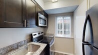 This single-floor residence offers a well-organized layout with a thoughtful mix of finishes throughout. The kitchen serves as a functional highlight, featuring dark espresso cabinetry with brushed gold hardware, granite countertops with subtle speckled detailing, a stainless steel undermount sink, and a full suite of stainless steel appliances including an electric range and over-the-range microwave. A window with vertical blinds allows natural light to brighten the space while offering outdoor views.

The living areas are generously proportioned, finished with neutral-toned carpet and painted in warm greige tones that create a calm, cohesive atmosphere throughout. A hallway connects the main living space to the bedroom areas, with hardwood-style flooring transitioning at the entryway. One bedroom notably features two side-by-side closets, providing ample storage with built-in hanging rods.

The bathroom is cleanly appointed with a dark vanity cabinet topped with a granite countertop, a mirror with an elegant multi-bulb light fixture above, and wood-look flooring. A bathtub with a surrounding surround completes the space. Overall, the unit presents a clean, move-in ready environment with consistent, updated finishes.