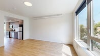 This well-designed single-floor unit offers a clean, modern aesthetic throughout, characterized by crisp white walls, light wood-tone flooring, and an abundance of natural light streaming through large windows. The open-plan layout seamlessly integrates the kitchen and living area, maximizing the efficient use of space. The kitchen is outfitted with warm wood-grain cabinetry, light-toned countertops, and a full suite of stainless steel appliances — including a refrigerator, electric range, over-the-range microwave, dishwasher, and a single-basin sink — creating a functional and stylish cooking environment. Recessed lighting runs along the kitchen corridor, complementing the natural light that filters in from the windows framing views of surrounding greenery. The living area is open and versatile, with large windows along one wall allowing sunlight to pour across the hardwood-style floors. The bathroom features a wood-tone vanity with an integrated sink, a large mirror, and a full shower-tub combination, all finished in clean, neutral tones. Throughout the unit, the design prioritizes simplicity and practicality, with consistent flooring, neutral color palettes, and modern fixtures that create a cohesive and inviting atmosphere.