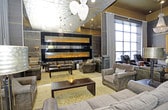 This striking single-floor space makes an immediate impression with its soaring ceilings and opulent Art Deco-inspired design. The main lounge area is anchored by a sleek linear fireplace set into a dark, richly textured wall, flanked by large floor-to-ceiling windows that flood the space with natural light and are dressed with flowing floor-length drapes. The ceiling is a standout architectural feature, showcasing a distressed metallic finish with gold accents that lend a sense of aged grandeur and visual depth. Pendant lighting with delicate globe fixtures cascades from above, complementing the warm glow of ornate table lamps with metallic shades. The furnishings are curated with intention — tufted leather seating, a nailhead-trimmed desk, and plush upholstered sofas in muted charcoal tones are arranged to create both intimate conversation areas and open circulation. The light herringbone-patterned flooring provides a refined contrast to the deep, moody palette of the walls and cabinetry. Dark lacquered cabinets and accent tables with geometric detailing reinforce the sophisticated aesthetic throughout, resulting in an environment that is at once dramatic and inviting.