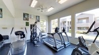 This fitness center is designed to support a well-rounded workout routine in a bright, functional environment. The space is outfitted with multiple cardio machines, including treadmills and an elliptical, alongside a multi-station cable and weight machine for comprehensive strength training. Large windows line the walls, flooding the room with natural light and providing views of the surrounding outdoor area, including landscaped grounds and adjacent buildings. The flooring consists of a durable dark rubberized surface, well-suited for high-impact activity and equipment placement. A ceiling fan is mounted overhead, contributing to ventilation and comfort during workouts. The walls are finished in a neutral tone with a textured ceiling, and informational fitness charts and posters are displayed, adding a practical, health-focused character to the space. The overall layout is efficient and open, allowing for easy movement between equipment. The single-floor facility offers a clean, accessible environment that caters to both cardiovascular and strength-training needs, making it a convenient and fully functional amenity for its users.