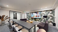 Located on W 41st Ave in Denver, CO, this thoughtfully designed lounge space offers a compelling blend of comfort and functionality within a single, well-appointed room. The space is anchored by a dramatic botanical-print accent wall that serves as a bold visual focal point, paired with a large multi-panel video display wall — ideal for presentations, media viewing, or casual entertainment.

The layout is divided into distinct zones to support a variety of uses. A central lounge area features modular tufted seating in deep charcoal and soft gray upholstery, arranged around a round wood coffee table adorned with decorative objects and greenery. A long communal table with bar-height stools along one side provides a dedicated workspace or dining area, offering flexibility for collaborative use.

On the opposite end, a light gray sectional sofa with colorful accent pillows faces a counter-height bar table lined with stools, beneath a curated gallery wall of framed abstract artwork. A sculptural dried botanical arrangement in a tall ceramic vessel adds an organic, artistic touch to the corner.

Recessed lighting and wall-mounted sconces cast a warm, even glow throughout, complementing the neutral carpet and clean white walls. Glass-paneled interior doors lend a sense of openness while maintaining defined boundaries. The overall ambiance is modern, polished, and inviting.