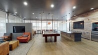 This thoughtfully designed single-floor amenity lounge blends modern industrial aesthetics with warm, inviting materials throughout. The main gathering area is anchored by a full-size billiards table set beneath elegant pendant lighting, with wide-plank hardwood flooring extending across the open layout. Floor-to-ceiling glass walls flood the space with natural light and offer direct views of an outdoor pool and patio area, creating a seamless indoor-outdoor connection enhanced by sheer roller shades for adjustable privacy.

A central bar and kitchenette features a reclaimed wood accent wall, a granite-topped island with an integrated sink, and Edison bulb pendant lighting overhead, with a mounted flat-screen TV completing the entertaining hub. Adjacent lounge seating areas are furnished with leather sofas, upholstered accent chairs, and rustic wood coffee tables, providing relaxed spaces for socializing or recreation, including tabletop games.

A separate dining and collaborative zone offers semi-private booth seating framed by teal structural columns and reclaimed wood-paneled walls, creating an intimate atmosphere ideal for small group gatherings. Dark tile flooring and moody overhead lighting give this area a distinctly cozy character. Throughout the space, the dark ceiling treatment, mixed material finishes, and carefully curated furnishings contribute to a cohesive, upscale ambiance.