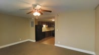 This single-floor space offers a practical and comfortable layout designed for everyday living. The living area features wall-to-wall carpeting in a warm neutral tone, creating a cozy atmosphere throughout. A ceiling fan with an integrated light fixture is centrally mounted overhead, providing both air circulation and ambient lighting to the room. The walls are finished in a soft beige tone, contributing to the overall warmth of the space.

Directly accessible from the living area is a well-appointed kitchen, visible through an open passageway that promotes a connected, open-concept feel. The kitchen is outfitted with rich, dark wood cabinetry that extends to the upper walls, offering generous storage. A black refrigerator and matching black range anchor the space, complemented by under-cabinet lighting that illuminates the countertops below. A sink is centrally positioned along the counter, adding to the kitchen's functional layout.

The transition between the carpeted living area and the hard-surface kitchen flooring is clearly defined, offering both visual contrast and practical distinction between spaces. An electrical panel is mounted on the living room wall, and recessed details in the ceiling add subtle architectural interest. Overall, the space presents a clean, functional environment suited for comfortable day-to-day use.