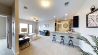 This single-floor residence is designed around an open-concept layout that unifies the living and kitchen areas into one cohesive, spacious environment. The living area is anchored by plush carpeting, a ceiling fan, and a large window that draws in natural light, creating a bright and comfortable atmosphere. A leather sofa, area rug, and coffee table furnish the space thoughtfully, while wall décor adds a subtle stylistic touch.

The kitchen is positioned along the far wall and features dark wood cabinetry, granite-look countertops, and stainless steel appliances, including a refrigerator and microwave. A wide peninsula counter with an integrated sink separates the kitchen from the living space, and three industrial-style bar stools provide casual seating along the breakfast bar. Recessed lighting illuminates the kitchen area effectively.

A hallway visible beyond the living area suggests additional rooms toward the rear of the unit. The textured ceiling, neutral wall tones, and consistent flooring throughout contribute to a clean, modern aesthetic that feels both functional and welcoming.