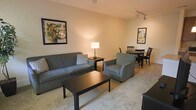 This single-floor space showcases an open-concept design that integrates the living and dining areas into one cohesive, functional layout. The living area is anchored by a plush gray sofa paired with a matching armchair, both arranged around a dark wood coffee table that rests atop soft, light-colored carpeting. A side table with a warm lamp adds ambient lighting to complement the track lighting mounted on the ceiling, which provides additional illumination throughout the space. Framed artwork on the neutral-toned walls adds character and visual interest to the room. The dining area is positioned adjacent to the living space, featuring a round dark wood table surrounded by matching chairs, making it an ideal spot for casual meals or gatherings. The open layout creates a natural flow between the two areas, contributing to an overall sense of spaciousness. The warm, neutral color palette used throughout — from the walls to the flooring — lends the space a calm and inviting ambiance, while the combination of ceiling and accent lighting ensures the interior remains bright and comfortable at any time of day.