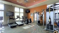 This fitness center is designed to provide a comprehensive workout experience within a thoughtfully appointed, single-floor space. The room features bold orange accent walls that lend an energizing ambiance, complemented by a tray ceiling with recessed lighting and a warm accent border that adds a polished architectural touch. Large windows along one wall flood the space with natural light, creating an open and inviting environment. The flooring is a durable, patterned carpet with orange geometric accents that tie into the overall color scheme. The facility is equipped with a range of modern cardio machines, including treadmills and ellipticals, as well as a multi-functional cable strength training tower, offering users a versatile full-body workout. A wall-mounted flat-screen television is positioned for easy viewing during exercise. A glass-paneled door provides access to an adjacent area, contributing to the sense of connectivity and flow. The space is well-organized and uncluttered, making it practical and easy to navigate. Overall, this fitness center combines functional equipment with an aesthetically cohesive design, delivering a motivating and comfortable environment for exercise.