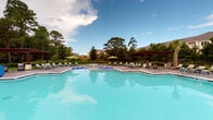Located at 3510 Corder Dr in Wilmington, North Carolina, this single-level outdoor amenity space is designed to provide a resort-style experience for its residents. At the heart of the space is a generously sized swimming pool with crystal-clear blue water, surrounded by an expansive pool deck lined with lounge chairs and shade umbrellas. A handsome wooden pergola anchors one end of the pool area, offering a shaded retreat with a refined aesthetic.

Adjacent to the pool, a covered outdoor lounge area features white structural columns and wraps around to provide open views of both the pool and the surrounding lush, tree-lined landscape. Comfortable modular seating in vibrant green adds a casual, welcoming touch to this transitional space. Black iron fencing borders the perimeter, defining the area while preserving the scenic views of mature trees and manicured landscaping.

The outdoor kitchen area is a standout feature, equipped with dual stainless steel grilling stations set against a white brick base and topped with a sleek dark granite countertop. A pass-through window connects the interior to the exterior entertaining space, enhancing functionality. Palm trees accent the grounds, reinforcing the coastal Carolina atmosphere. Overall, this space offers an inviting and well-appointed outdoor environment ideal for relaxation and social gatherings.