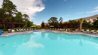 Located at 3510 Corder Dr in Wilmington, North Carolina, this single-level outdoor amenity space is designed to provide a resort-style experience for its residents. At the heart of the space is a generously sized swimming pool with crystal-clear blue water, surrounded by an expansive pool deck lined with lounge chairs and shade umbrellas. A handsome wooden pergola anchors one end of the pool area, offering a shaded retreat with a refined aesthetic.

Adjacent to the pool, a covered outdoor lounge area features white structural columns and wraps around to provide open views of both the pool and the surrounding lush, tree-lined landscape. Comfortable modular seating in vibrant green adds a casual, welcoming touch to this transitional space. Black iron fencing borders the perimeter, defining the area while preserving the scenic views of mature trees and manicured landscaping.

The outdoor kitchen area is a standout feature, equipped with dual stainless steel grilling stations set against a white brick base and topped with a sleek dark granite countertop. A pass-through window connects the interior to the exterior entertaining space, enhancing functionality. Palm trees accent the grounds, reinforcing the coastal Carolina atmosphere. Overall, this space offers an inviting and well-appointed outdoor environment ideal for relaxation and social gatherings.