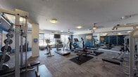 This fitness center is a comprehensively equipped facility designed to support a wide range of workout routines. The space is defined by a bold navy blue accent wall that anchors the room visually, complemented by neutral-toned carpeting and a textured ceiling overhead. Ceiling fans and flush-mount light fixtures are distributed throughout, ensuring comfortable airflow and consistent illumination.

The cardio zone features multiple treadmills, elliptical machines, and upright cycling equipment, all positioned to take advantage of the natural light streaming in through large windows that offer views of the surrounding exterior. A wall-mounted flat-screen television is positioned nearby for entertainment during workouts.

The strength training area includes a cable crossover machine, a multi-function weight rack, adjustable benches, and a squat/power rack station, providing a thorough selection of resistance training options. A dedicated free weight section houses a rack of dumbbells in varying sizes, accompanied by stability balls for functional training.

Full-length mirrors line one wall, enhancing the sense of openness while allowing users to monitor their form. A posted fitness center rules sign and a wall clock complete the practical details of the space. The overall ambiance is functional and well-organized, offering a focused environment suited for a variety of fitness goals.