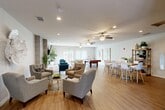 Located at 1610 Jenkins Rd in Bakersfield, California, this thoughtfully designed single-floor community clubroom offers a welcoming, open-plan layout that accommodates a variety of social and recreational activities. The space flows seamlessly across light-toned hardwood-style flooring, unified by a clean, contemporary aesthetic and a neutral color palette of soft whites and warm grays.

The lounge area serves as the heart of the space, anchored by a floor-to-ceiling tile-clad fireplace topped with a large wall-mounted flat-screen TV. Comfortable gray tufted armchairs and vibrant teal seating are arranged to encourage conversation, complemented by a decorative white sculptural wall piece that adds an artistic focal point. Recessed lighting and dual ceiling fans maintain a bright, comfortable atmosphere throughout.

Adjacent to the lounge, a game area features a full-size pool table, while a dining zone offers a live-edge wood table surrounded by modern white chairs, ideal for group gatherings. Natural light streams in through large sliding glass doors that open to outdoor views, enhancing the airy feel of the space.

The kitchenette is well-appointed with dark espresso cabinetry, a classic white subway tile backsplash, light countertops, and stainless steel appliances including a refrigerator and dishwasher, making it fully functional for hosting events.