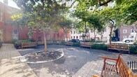 Located at 200 West Washington Square in Philadelphia's historic Old City neighborhood, this outdoor courtyard space offers a thoughtfully designed urban retreat that seamlessly merges classic architecture with contemporary landscaping. The space is anchored by a striking red brick building featuring tall, arched windows with ornate ironwork detailing, characteristic of Philadelphia's rich architectural heritage. A modern multi-story building provides a contrasting backdrop, highlighting the dynamic blend of historic and contemporary design. Brick-paved walkways wind through the courtyard, flanked by curated garden beds filled with ferns, flowering shrubs, ornamental grasses, and mature trees that provide generous shade and greenery. Stone edging and black wrought iron fencing define the garden borders, adding a refined, classic touch throughout. Outdoor seating areas, complete with tables shaded by dark market umbrellas and wooden benches, invite visitors to linger and enjoy the open-air environment. Large-format concrete pavers create generous open areas suitable for vehicle access or gathering. The overall ambiance is one of a well-maintained, serene urban oasis that balances the energy of the surrounding city with thoughtful green design, making it an appealing and functional exterior space in the heart of Philadelphia.