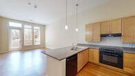 This well-designed one-bedroom, one-bathroom residence in Denver, Colorado offers a thoughtfully organized single-floor layout. The open-concept kitchen and living area serves as the heart of the home, featuring warm hardwood flooring, high ceilings, and large windows with a glass door that invite abundant natural light and provide views of the surrounding outdoor greenery. The kitchen is equipped with light maple cabinetry, granite countertops, a peninsula with a double sink, pendant lighting, and black appliances including a gas range and refrigerator — offering both functionality and style. The living area is generously proportioned and flows seamlessly from the kitchen. The bedroom features plush carpeting, neutral warm-toned walls, and a window with blinds that allows for natural light. The full bathroom includes tile flooring, a wood-toned vanity with a dark countertop, accessibility grab bars, and a tiled shower/tub combination. A dedicated laundry room houses a full-size front-loading washer and dryer alongside built-in shelving and additional cabinetry, adding notable convenience. The space maintains a clean, neutral palette throughout, creating a bright and versatile living environment.