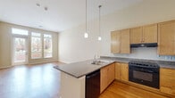 This well-designed one-bedroom, one-bathroom residence in Denver, Colorado offers a thoughtfully organized single-floor layout. The open-concept kitchen and living area serves as the heart of the home, featuring warm hardwood flooring, high ceilings, and large windows with a glass door that invite abundant natural light and provide views of the surrounding outdoor greenery. The kitchen is equipped with light maple cabinetry, granite countertops, a peninsula with a double sink, pendant lighting, and black appliances including a gas range and refrigerator — offering both functionality and style. The living area is generously proportioned and flows seamlessly from the kitchen. The bedroom features plush carpeting, neutral warm-toned walls, and a window with blinds that allows for natural light. The full bathroom includes tile flooring, a wood-toned vanity with a dark countertop, accessibility grab bars, and a tiled shower/tub combination. A dedicated laundry room houses a full-size front-loading washer and dryer alongside built-in shelving and additional cabinetry, adding notable convenience. The space maintains a clean, neutral palette throughout, creating a bright and versatile living environment.