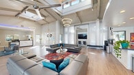 Located on U.S. 98 in Pensacola, Florida, this single-floor commercial space delivers a striking blend of industrial and contemporary design. The main living and lounge area commands attention with dramatically tall ceilings supported by exposed structural beams and trusses, while tiered ring pendant lights and ceiling fans hang overhead. Floor-to-ceiling windows and sets of glass-paned French doors line the walls, flooding the interior with natural light and offering views of the outdoor surroundings. Light hardwood flooring runs throughout, unifying the open layout. Plush gray leather sectional sofas are arranged around a vibrant, multicolored area rug, creating a welcoming and stylish lounge atmosphere. A wall-mounted flat-screen TV and built-in workstation nook with sleek office chairs provide functional workspace alongside a comfortable lounge bench. The reception area near the entrance features a marble-topped desk, modern accent chairs on a teal rug, and a sense of openness accentuated by vaulted ceilings. The galley-style kitchen is well-appointed with white shaker cabinetry, dark countertops, stainless steel fixtures, and ample workspace on both sides. A café bar area, branded as the Cafe Bar of Nottingham, adds a unique hospitality element with a professional coffee station and prep counter, making this space as functional as it is visually impressive.