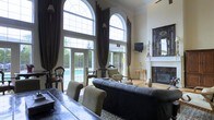 This sophisticated single-floor space makes an immediate impression with its soaring double-height ceilings and a striking pair of grand arched windows crowned with elegant transom detailing. Floor-to-ceiling dark chocolate drapes frame the windows and French doors, adding a sense of drama while controlling natural light that floods the interior. The living area is anchored by a tufted black leather sofa and complemented by traditional wooden armchairs with upholstered cushions, all arranged atop a patterned area rug. A classic white fireplace mantel with an ornate dark-framed mirror above it serves as a refined focal point along one wall. A decorative console table with slender gold legs adds an antique accent nearby. The French doors open directly to an outdoor pool area, seamlessly connecting the interior to the exterior and enhancing the sense of openness. A wall-mounted flat-screen television is positioned above the fireplace area, balancing modern convenience with the room's traditional aesthetic. The overall ambiance is one of formal elegance, combining classic architectural details with rich furnishings to create a stately and welcoming atmosphere.