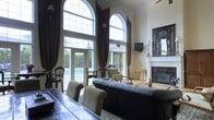 This sophisticated single-floor space makes an immediate impression with its soaring double-height ceilings and a striking pair of grand arched windows crowned with elegant transom detailing. Floor-to-ceiling dark chocolate drapes frame the windows and French doors, adding a sense of drama while controlling natural light that floods the interior. The living area is anchored by a tufted black leather sofa and complemented by traditional wooden armchairs with upholstered cushions, all arranged atop a patterned area rug. A classic white fireplace mantel with an ornate dark-framed mirror above it serves as a refined focal point along one wall. A decorative console table with slender gold legs adds an antique accent nearby. The French doors open directly to an outdoor pool area, seamlessly connecting the interior to the exterior and enhancing the sense of openness. A wall-mounted flat-screen television is positioned above the fireplace area, balancing modern convenience with the room's traditional aesthetic. The overall ambiance is one of formal elegance, combining classic architectural details with rich furnishings to create a stately and welcoming atmosphere.