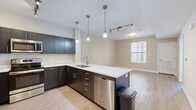 Located at 11901 S Freedom Park Dr in Herriman, Utah, this single-floor unit offers a thoughtfully designed layout with modern finishes throughout. The kitchen serves as a central highlight, showcasing dark espresso cabinetry, crisp white quartz countertops, and a full suite of stainless steel appliances including a refrigerator, microwave, and range. A peninsula with an integrated sink extends the workspace and provides casual seating potential, while a combination of pendant lights and track lighting ensures excellent illumination. Light-toned wood-look flooring flows through the kitchen and extends into the adjoining living areas, creating a seamless, cohesive feel. The living space is bright and open, benefiting from natural light admitted through blinds-equipped windows. Bedrooms are generously proportioned, with some featuring plush carpet flooring and large windows that invite natural light. A closet with louvered doors adds practical storage in one of the bedrooms. The bathroom is finished with large-format tile flooring, white cabinetry with brushed nickel hardware, a wood-tone countertop, a framed mirror, and a full soaking tub with shower. Neutral warm-toned walls throughout the unit create a calm, welcoming ambiance well-suited for comfortable everyday living.