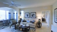 This single-floor residence showcases a thoughtfully designed living area that balances style and comfort. The living room serves as the centerpiece of the space, featuring neutral beige walls that provide a warm, versatile backdrop. A ceiling fan overhead ensures year-round comfort while contributing to the room's understated elegance.

Natural light flows generously through multiple windows dressed with layered curtains and blinds, creating a bright yet controllable ambiance. The room is anchored by a plush, cream-colored sofa accented with blue and navy throw pillows, complemented by a patterned accent chair in muted green tones. A dark-finished, circular coffee table anchors the seating arrangement atop a decorative area rug with a medallion pattern.

Two framed black-and-white artworks are displayed symmetrically on the main wall, adding a refined, contemporary touch. Matching table lamps on dark wood end tables provide warm task lighting. A graceful arched doorway connects the living area to adjacent spaces, enhancing the open and cohesive flow throughout the home. Light-toned tile flooring ties the rooms together seamlessly.