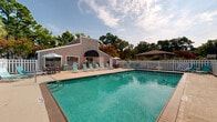 Located at 200 Branchwood Dr in Goose Creek, South Carolina, this outdoor community pool area offers a well-appointed recreational space designed for relaxation and leisure. The centerpiece is a large, rectangular swimming pool with clear turquoise water, accessible via stainless steel entry rails on multiple sides. The expansive concrete and wood-decked surround provides ample room for sunbathing and socializing, furnished with numerous teal lounge chairs positioned along the pool's perimeter. A dedicated poolside structure serves as a clubhouse or amenity building, featuring large windows, a distinctive navy awning, and an adjacent patio area outfitted with umbrella-shaded tables and chairs — ideal for outdoor dining or gatherings. The entire pool area is enclosed by a white vinyl privacy fence, providing both security and a clean, cohesive aesthetic. Lush mature trees, including palms and oaks, frame the surroundings, offering natural shade and a pleasant green backdrop. The layout, as seen from an aerial perspective, reveals a thoughtfully organized space that balances open swimming area with comfortable lounging zones. Under the open South Carolina sky, this community pool area presents an inviting outdoor retreat well-suited for warm-weather enjoyment.