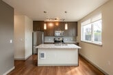 This single-floor residence offers a thoughtfully designed layout with a clean, contemporary aesthetic throughout. A welcoming hallway with rich hardwood flooring and recessed lighting leads seamlessly into the main living areas, creating a bright and cohesive flow from entry to living space.

The kitchen is a standout feature, showcasing rich dark wood cabinetry extending to the ceiling for generous storage, stainless steel appliances including a refrigerator, electric range, and microwave, complemented by a textured tile backsplash. A peninsula with a granite-style countertop and a built-in sink doubles as a prep area and casual dining space, with pendant lighting adding warmth overhead. Natural light filters in through a nearby window, enhancing the kitchen's open feel.

The bathroom features warm hardwood flooring, a dark wood vanity with a granite countertop, a large mirror, and a full soaking tub with a tiled surround. A second bathroom mirrors this refined style, offering a similarly appointed vanity and a well-lit, functional layout.

Additional rooms feature soft carpeting, neutral wall tones, and ample natural light, providing versatile spaces suited to a variety of uses. Consistent use of hardwood flooring, neutral palettes, and modern hardware throughout the residence ties each area together into a cohesive and polished whole.