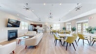 Located at 3701 Lilac Ln in Enola, PA, this community clubhouse spans two floors and offers a well-appointed gathering space with a bright, modern aesthetic. The main floor features an open-concept layout that seamlessly connects the living room, dining area, and kitchen. Soaring vaulted ceilings with recessed lighting and stylish sputnik-style chandeliers create a refined yet welcoming ambiance throughout. Wide-plank light hardwood floors run continuously across the space, enhancing its airy feel. The living area is anchored by a gas fireplace and complemented by cream-toned sofas, accent chairs, and vibrant abstract wall art. A wall-mounted television adds a contemporary touch. The dining area accommodates multiple seating arrangements with modern chartreuse chairs and white square tables, bathed in natural light from large windows and glass doors. The fully equipped kitchen features rich dark-wood cabinetry, stainless steel appliances, a classic white subway tile backsplash, and a marble-topped island with bar seating. A welcoming entry vestibule with double glass doors and coat storage completes the interior. Outdoors, a large swimming pool with a surrounding sundeck, lounge chairs, and an upper-level deck offers a resort-like setting ideal for community gatherings.
