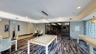 Located at 4800 Marriott Dr in Raleigh, North Carolina, this single-floor amenity space is designed to offer both recreation and social gathering opportunities. The main area is anchored by a contemporary pool table with a sleek white and wood finish, positioned beneath a modern rectangular pendant light fixture that provides focused illumination. The flooring features a bold, multi-toned patterned carpet that adds visual depth and energy to the space. Recessed lighting throughout the ceiling complements the pendant fixture, ensuring the area is well-lit and inviting. A raised tray ceiling detail adds an element of architectural refinement to the room. A bistro-style high-top table with white chairs is tucked into one corner, offering a casual spot for conversation or refreshments. Adjoining the main recreation area is a semi-private lounge or meeting room, separated by elegant glass bi-fold or accordion-style doors with dark metal framing. This secondary space is furnished with upholstered chairs and features a mounted flat-screen display, making it suitable for meetings, presentations, or media viewing. The overall ambiance is polished and modern, blending functional amenities with contemporary interior design elements.