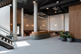 This striking commercial lobby, operating under the Fleethouse brand, presents an exceptional blend of industrial and contemporary design. The entrance immediately impresses with its soaring double-height ceilings, floor-to-ceiling windows, and a dramatic staircase featuring glass railings and rich wood treads, flooding the space with abundant natural light.

The main lobby is anchored by a curved, sage-green reception desk set against a stunning vertical-slat wood accent wall, which serves as a dramatic architectural backdrop alongside a large textile art installation. Polished light grey tile flooring extends throughout, complementing the exposed concrete columns and industrial-style exposed ductwork overhead.

Multiple distinct lounge areas offer varied seating experiences. A curved navy velvet banquette with a sculptural wooden side table sits beneath pendant lighting near the entrance. A warm-toned leather banquette seating area with channel-tufted detailing, brass accent lamps, and marble-topped tables provides an intimate gathering nook. Additional seating includes rich terracotta velvet armchairs and a fireplace-adjacent lounge with red upholstered booth seating.

Throughout, greenery in contemporary planters adds life, while gold-toned pendant lights and wall sconces layer warmth into the otherwise refined, neutral palette — creating a lobby that is both functional and design-forward.