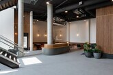 This striking commercial lobby, operating under the Fleethouse brand, presents an exceptional blend of industrial and contemporary design. The entrance immediately impresses with its soaring double-height ceilings, floor-to-ceiling windows, and a dramatic staircase featuring glass railings and rich wood treads, flooding the space with abundant natural light.

The main lobby is anchored by a curved, sage-green reception desk set against a stunning vertical-slat wood accent wall, which serves as a dramatic architectural backdrop alongside a large textile art installation. Polished light grey tile flooring extends throughout, complementing the exposed concrete columns and industrial-style exposed ductwork overhead.

Multiple distinct lounge areas offer varied seating experiences. A curved navy velvet banquette with a sculptural wooden side table sits beneath pendant lighting near the entrance. A warm-toned leather banquette seating area with channel-tufted detailing, brass accent lamps, and marble-topped tables provides an intimate gathering nook. Additional seating includes rich terracotta velvet armchairs and a fireplace-adjacent lounge with red upholstered booth seating.

Throughout, greenery in contemporary planters adds life, while gold-toned pendant lights and wall sconces layer warmth into the otherwise refined, neutral palette — creating a lobby that is both functional and design-forward.