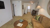 This single-floor residence offers a comfortable, open layout that seamlessly connects the living, dining, and entry areas. The living room is carpeted in warm neutral tones and centers around a round wooden coffee table paired with a plush sofa accented by green throw pillows, creating an inviting atmosphere. A ceiling fan with a light fixture overhead provides both ventilation and ambient lighting, while a tall floor lamp adds a warm glow to the corner.

Adjacent to the living area, a compact dining nook accommodates a dark wood bistro-style table and chairs, ideal for casual meals. The space flows naturally toward a private entrance door at the rear, which allows natural light to filter into the unit, brightening the interior. A stainless steel refrigerator is visible near the entry corridor, hinting at a kitchen tucked efficiently within the layout.

Storage is well-addressed, with a louvered closet door and an additional open closet visible along one wall, offering practical organization options. The neutral wall tones and carpeted flooring throughout contribute to a cohesive, understated aesthetic suited for everyday living.