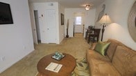 This single-floor residence offers a comfortable, open layout that seamlessly connects the living, dining, and entry areas. The living room is carpeted in warm neutral tones and centers around a round wooden coffee table paired with a plush sofa accented by green throw pillows, creating an inviting atmosphere. A ceiling fan with a light fixture overhead provides both ventilation and ambient lighting, while a tall floor lamp adds a warm glow to the corner.

Adjacent to the living area, a compact dining nook accommodates a dark wood bistro-style table and chairs, ideal for casual meals. The space flows naturally toward a private entrance door at the rear, which allows natural light to filter into the unit, brightening the interior. A stainless steel refrigerator is visible near the entry corridor, hinting at a kitchen tucked efficiently within the layout.

Storage is well-addressed, with a louvered closet door and an additional open closet visible along one wall, offering practical organization options. The neutral wall tones and carpeted flooring throughout contribute to a cohesive, understated aesthetic suited for everyday living.