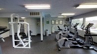 This single-floor fitness center provides a functional and well-organized workout environment designed to accommodate a range of exercise needs. The space features durable rubber flooring throughout, offering both safety and comfort underfoot during workouts. Bright fluorescent overhead lighting ensures the entire area is well illuminated, creating a practical and focused atmosphere for exercise.

The cardio section is equipped with multiple elliptical machines and stationary bikes, positioned near a window that allows natural light to filter in and provides a view of the surrounding greenery. A wall-mounted television is conveniently positioned nearby, offering entertainment during workouts. Framed informational posters and notices are displayed along the walls, adding a structured, facility-oriented feel to the space.

On the strength training side, a weight machine is available for resistance exercises, catering to users looking to build and tone muscle. The room also includes a dedicated entry area with lockers for personal storage, enhancing the usability of the space. HVAC venting is visible near the ceiling, ensuring proper climate control throughout the facility. A waste bin is conveniently placed within the main workout area. Overall, this fitness center delivers a straightforward, no-frills exercise environment with all the essential equipment needed for a well-rounded workout routine.