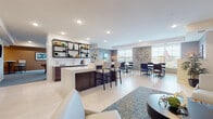 This single-floor community amenity space is thoughtfully designed to accommodate a variety of social activities, combining several distinct zones within a cohesive, contemporary layout. Light-toned flooring flows throughout, unifying the space while recessed lighting ensures consistent illumination across each area.

The central wet bar serves as a focal point, featuring a dark wood base, a bright white countertop, and seating for several guests on modern bar stools. Adjacent open shelving with a subway tile backsplash provides both functional storage and decorative display. A lounge area nearby offers comfortable seating arranged around a round coffee table, complemented by a patterned area rug that anchors the space.

The dining zone is bathed in natural light from large windows and is accented by a striking oversized clock on a textured gray accent wall, with multiple tables and upholstered chairs arranged for group gatherings. A dedicated TV lounge area provides a relaxed setting with armchairs, framed artwork, and a wall-mounted television above a dark media console. A separate billiards room features a full-size pool table, patterned wallcovering, and bistro-style seating, offering a more recreational retreat within the overall space.