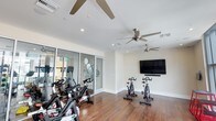 This single-floor fitness studio is thoughtfully designed to support an active lifestyle. The main workout area features rich hardwood flooring that adds warmth to the otherwise clean, contemporary aesthetic defined by crisp white walls and a bright, open layout. Multiple ceiling fans are strategically positioned throughout the space, ensuring consistent airflow during intense workout sessions. Recessed lighting complements the overall illumination, creating a well-lit environment suitable for exercise at any time of day.

A row of spin bikes is arranged along one side of the room, offering dedicated space for cycling workouts. A large wall-mounted flat-screen television, paired with a soundbar, is positioned at an optimal viewing angle for entertainment or guided workout sessions. The studio also includes a glass-walled adjacent room visible through floor-to-ceiling sliding glass panels, which appears to contain additional fitness equipment such as weight plates and other accessories. This transparent partition allows for visual connectivity between spaces while maintaining functional separation, adding a sense of depth and versatility to the overall fitness facility.