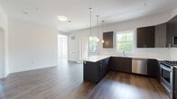 Located at 310 Madison Ave in Elizabeth, NJ, this single-floor residence showcases a thoughtfully designed open-concept layout with contemporary finishes throughout. The kitchen serves as the heart of the space, featuring dark espresso cabinetry, a large center island with a crisp white quartz countertop, stainless steel appliances including a gas range, built-in microwave, and refrigerator, all illuminated by sleek pendant lighting. Rich hardwood-style flooring flows seamlessly from the kitchen into the open living area, creating a warm and cohesive aesthetic. Multiple windows invite an abundance of natural light, while a set of glass sliding doors leads to a private balcony with a metal railing, offering outdoor views of surrounding greenery. The bedroom area is generously sized, with large windows that frame tree-lined views and bathe the room in sunlight. The bathroom is a standout feature, showcasing large-format marble-look tiles on both the floor and shower/tub surround, paired with a dark-wood vanity and a clean white countertop, exuding a refined, spa-like quality. The overall ambiance is modern, clean, and bright, making this a well-appointed and stylish living space.