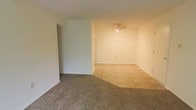 This single-floor space offers a clean, straightforward layout designed for practical everyday living. The main living area is a standout feature, showcasing a seamless transition between two distinct flooring types — warm, neutral-toned carpet and square tile — visually defining different functional zones within the open floor plan. The ceiling-mounted fan with integrated lighting provides both air circulation and ambient illumination, contributing to the room's comfortable atmosphere. The walls are finished in a clean, neutral white, offering a bright and versatile backdrop. Structural details such as a partial interior wall create a natural division within the space, adding architectural interest while maintaining an open feel. Electrical outlets are conveniently positioned along the baseboards, and the overall layout reflects a no-frills, functional design. The space is well-lit and presents a blank canvas that can accommodate a variety of furnishing arrangements and personal styles. Its straightforward, single-level design makes it an accessible and easy-to-navigate environment throughout.