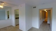 This single-floor residence offers a clean and functional layout designed for comfortable everyday living. The main living area features neutral-toned carpet flooring and light-painted walls, creating a bright and airy atmosphere throughout. A centrally located HVAC vent in the ceiling ensures efficient climate control across the space.

The kitchen, accessible directly from the main living area, is compact yet well-organized. It features white cabinetry with ample storage, a sink with a countertop workspace, and tile flooring that contrasts with the carpeted areas. The white cabinetry and clean lines give the kitchen a neat, practical aesthetic.

A hallway extending from the main living area leads to additional rooms, hinted at by the series of doors visible along its length. The hallway is carpeted in continuity with the living area, maintaining a cohesive flow throughout the space. The doors feature classic panel designs with brass-toned hardware, adding a modest traditional touch.

Overall, the space presents a straightforward and well-maintained interior with a consistent, neutral color palette of whites and beiges. The open sightlines between the living area and kitchen enhance the sense of spaciousness, making this a practical and inviting single-floor layout.